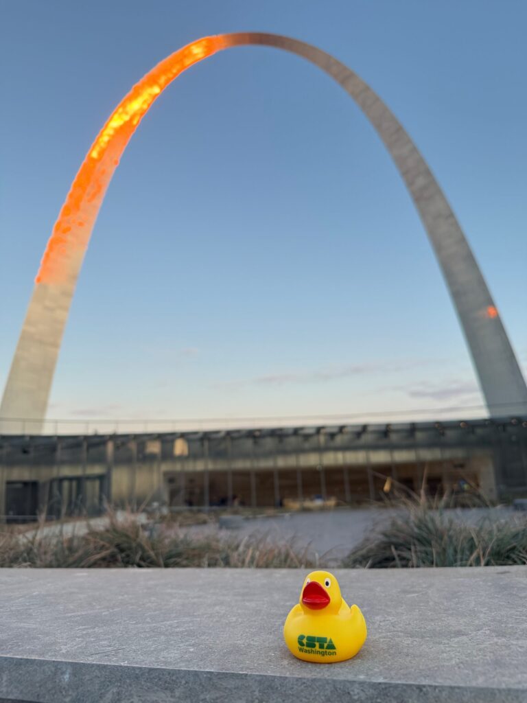 A yellow duck with the CSTA Washington logo on it sitting on a bench in front of the St. Louis arch lit up at sunset.